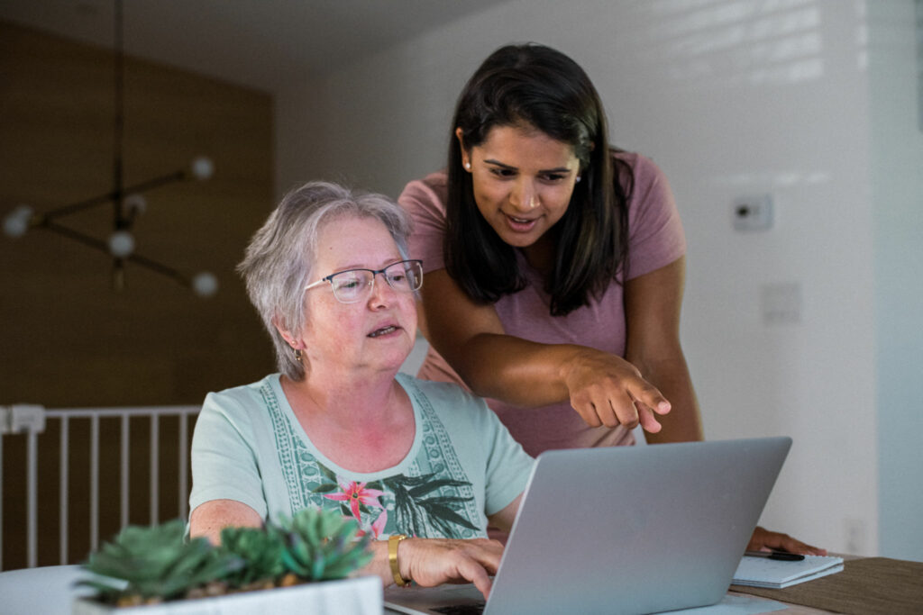 A Caucasian senior woman sits at a table in her home, working on a laptop computer as her multiracial adult daughter stands beside her, helping.